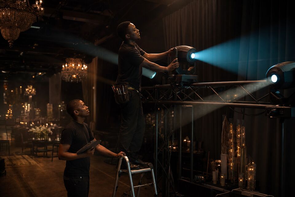 A lighting technician focusing a fixture on a low truss while soft cyan beams cut through light haze