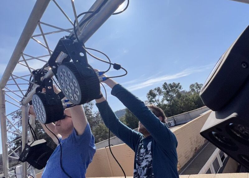 Two technicians rigging LED stage lights onto truss against a blue sky