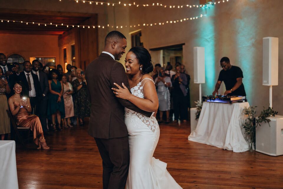 A newlywed couple's first dance under warm festoon lights, guests gathered around the dance floor